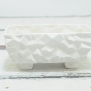 May include: White ceramic planter with a textured, geometric design. The planter has four short legs.