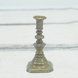 May include: A small, antique brass candlestick with a simple, elegant design. The candlestick has a round base and a tall, slender stem.