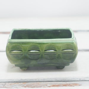 Puede incluir: Un macetero cuadrado de cerámica verde con un diseño en relieve de formas redondeadas. El macetero tiene tres pequeños pies en la parte inferior.
