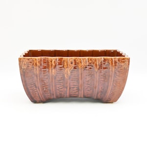 以下が含まれることがあります： リブ模様の長方形の茶色のセラミックプランター。 プランターは湾曲したベースと、上部に濃い茶色のテクスチャード加工と艶出し仕上げが施されています。 長さは約25cmです。