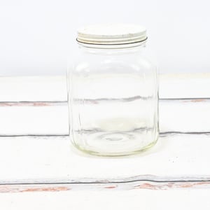 Puede incluir: Un tarro de vidrio transparente con una tapa blanca. El tarro tiene una superficie texturizada y está vacío.