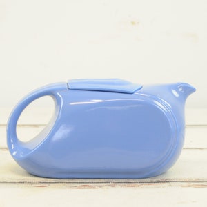 May include: A light blue ceramic teapot with a lid and a handle. The teapot is on a white wooden surface.