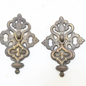 May include: Two matching antique brass drawer pulls featuring an ornate, symmetrical scrollwork backplate and a central drop handle. The metal has a darkened, aged patina with raised detailing and a small, decorative finial at the bottom of each piece.