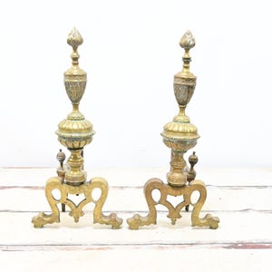 Puede incluir: Par de morillos de chimenea antiguos de latón y tono dorado. Cada morillo presenta un remate decorativo, un cuerpo central en forma de urna y una base en espiral. Los morillos se exhiben sobre un fondo blanco.