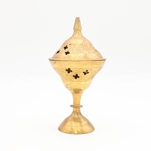 May include: A gold-colored incense burner with a conical lid and a flared base. The lid and body feature decorative cut-out patterns and etched designs. The burner stands on a pedestal base.