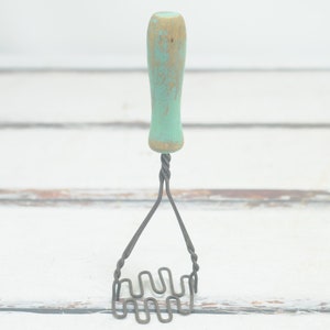 May include: A vintage wire potato masher with a light blue and yellow wooden handle. The masher is on a white wooden surface.