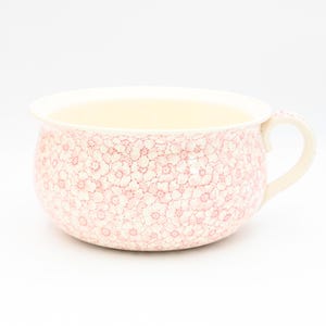 May include: A white ceramic chamber pot with a pink floral pattern. The pot has a single handle on the side.