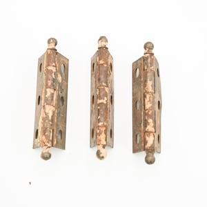 Puede incluir: Tres bisagras metálicas antiguas con un aspecto envejecido y desgastado. Cada bisagra es rectangular con múltiples orificios para tornillos y un remate decorativo en forma de bola en cada extremo. Las bisagras tienen un color marrón oxidado y desgastado con restos de pintura descolorida.