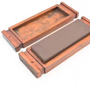 May include: A rectangular sharpening stone in a wooden box. The stone is a dark brown color and sits inside a wooden frame. The box is open, revealing the stone and its housing. The wood has a reddish-brown hue.