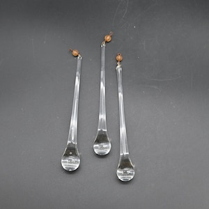May include: Three clear glass teardrop ornaments with small brown beads at the top. The ornaments are elongated with a bulbous bottom and hang against a dark grey background. These decorative glass ornaments are ideal for holiday decor.
