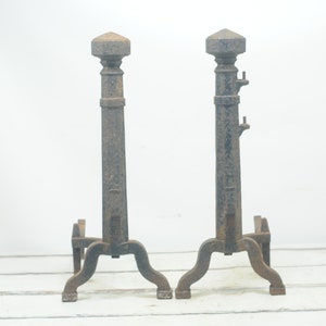 Puede incluir: Un par de soportes de chimenea antiguos de hierro fundido con un acabado gris oscuro. Los soportes tienen una parte superior decorativa y una base de tres patas.