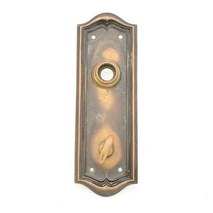 May include: A rectangular bronze door plate with a dark patina and gold-toned highlights. The plate features a central circular opening for a knob and a lower thumb turn mechanism. It has a decorative arched top and bottom with four screw holes.