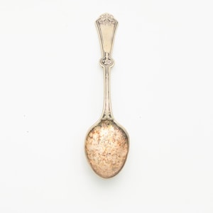 May include: A vintage silver spoon with an ornate handle and a tarnished bowl. The spoon has a decorative design at the top of the handle and a rounded bowl. The spoon is set against a white background.
