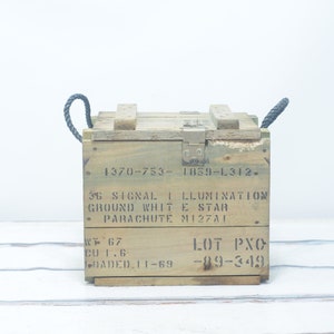 May include: A wooden box with rope handles and a metal latch. The box is labeled "36 SIGNAL ILLUMINATION GROUND WHITE STAR PARACHUTE M127A1" and "LOT PXO -89-349".