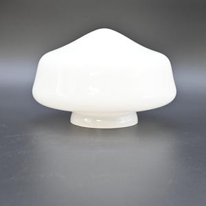 Könnte beinhalten: Ein weißer, pilzförmiger Lampenschirm aus Glas. Der Schirm hat eine abgerundete Oberseite und einen ausgestellten Sockel, der auf einer dunkelgrauen Oberfläche steht. Das opake Glas hat eine glänzende Oberfläche und reflektiert das Licht.