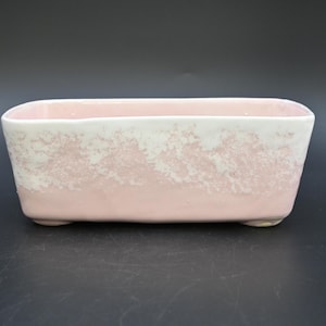May include: A rectangular ceramic planter with a pink base and white speckled top. The planter has rounded corners and sits on small feet. The background is a solid black.