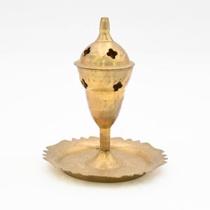 May include: A gold metal incense burner with a decorative design. The burner has a round base and a tall, conical top with holes for incense.
