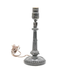May include: A dark grey antique-style table lamp with a decorative base and stem. The lamp has a standard bulb socket and a brown electrical cord. The lamp is designed to sit on a table or desk and provide light.