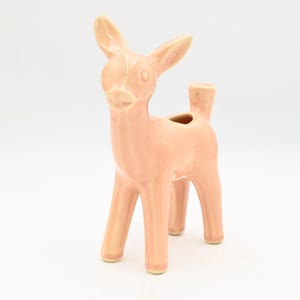 Puede incluir: Una figura de ciervo de cerámica en color melocotón suave. El ciervo tiene un cuerpo esbelto, cuatro patas y una pequeña abertura en la espalda. Las orejas están erguidas y el ciervo está sobre una superficie blanca.