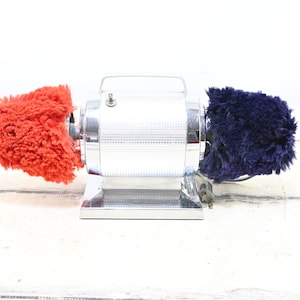 Puede incluir: Un pulidor de zapatos vintage con un acabado cromado brillante. El pulidor tiene un cuerpo cilíndrico con un asa y dos almohadillas de pulido esponjosas, una roja y otra azul marino. El dispositivo está diseñado para pulir zapatos.