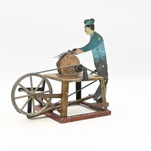 Puede incluir: Un juguete de madera vintage que representa a un hombre afilando una cuchilla en una muela. La figura está vestida de azul y verde, con una rueda de madera y una base roja. El juguete es probablemente de principios del siglo XX.