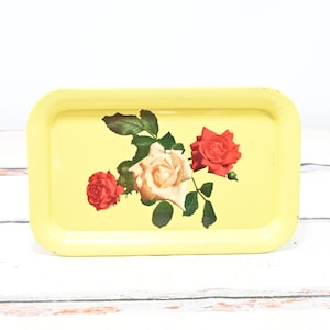 May include: A rectangular yellow metal tray with rounded corners. The tray features a floral design of red and cream roses with green leaves. The tray is set against a white and wood-grain background.