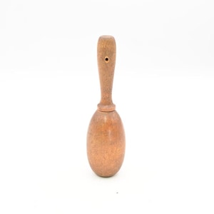 Puede incluir: Una maraca de madera marrón con un cuerpo liso de forma ovalada y un mango cilíndrico delgado. El mango incluye un pequeño orificio circular cerca de la parte superior, y todo el instrumento muestra una textura natural de veta de madera.