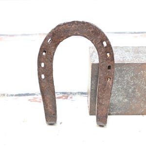 May include: A rusty, antique horseshoe with visible nail holes, standing upright next to a square, textured metal block. The horseshoe is a dark brown color, suggesting age and use. The background is a white surface.