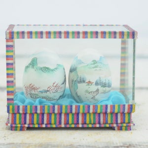 Puede incluir: Dos esferas de vidrio blancas con paisajes pintados dentro de una caja de rayas coloridas con tapa de vidrio. Las esferas están sobre una base de tela azul.