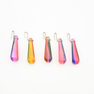 May include: Five iridescent crystal prism pendants with silver-colored hooks. The teardrop-shaped prisms display a gradient of colors, including pink, yellow, green, and blue. These decorative prisms are designed to hang and catch the light.
