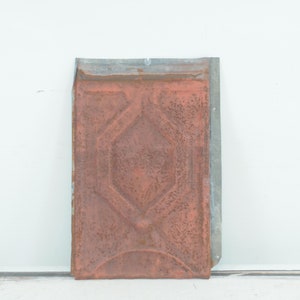 May include: A rusty metal tile with a geometric pattern. The tile has a raised design and is a reddish-brown color.