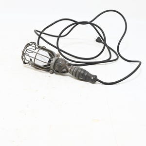 May include: A portable work light featuring a dark, ribbed handle and a protective metal wire cage surrounding the bulb housing. The light is connected to a long black electrical cord with a standard plug, designed for handheld illumination.