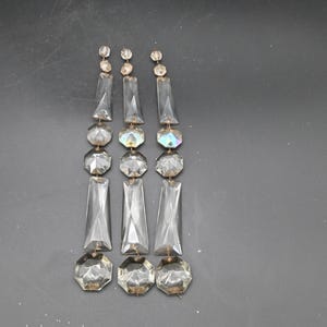 Op de afbeelding: Drie strengen heldere kristallen kroonluchterprismas. Elke streng heeft een reeks gefacetteerde, rechthoekige en ronde kristallen. De kristallen zijn verbonden door kleine metalen pinnen. De achtergrond is donkergrijs.