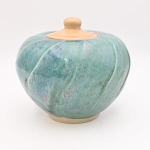 Op de afbeelding: Een keramische pot met een deksel. De pot heeft een afgeronde vorm met een turquoise en blauwe glazuur. Het deksel en de basis zijn lichtbeige van kleur. De pot heeft een gestructureerd, geribbeld ontwerp. De pot is waarschijnlijk handgemaakt.