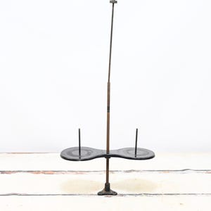 May include: An antique spool holder with a dark metal base and a two-tiered, oval-shaped platform. A tall, thin rod extends upwards, with a smaller platform at the top. The holder is designed to hold spools of thread.