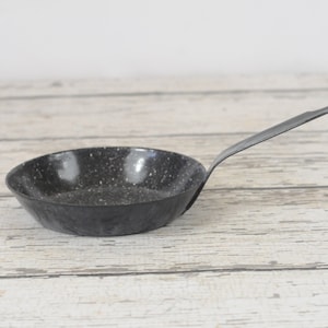 Puede incluir: Una sartén pequeña, negra y moteada con un mango negro. La sartén está sobre una superficie de madera blanca.