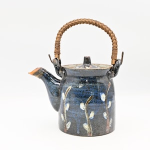 May include: A ceramic teapot with a dark blue and black glazed finish featuring a hand-painted pussy willow branch design. The teapot includes a matching lid and a curved, woven brown rattan handle attached to two metal loops on the sides.