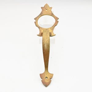 May include: Ornate brass door handle with a decorative design. The handle features a central grip with an open circle and decorative elements at the top and bottom. The brass has a warm, aged patina, suggesting vintage or antique origins.