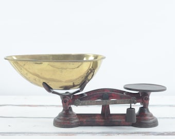 May include: Antique red and brass kitchen scale with a round weighing pan and a bowl-shaped pan. The scale has a black metal pointer and a brass weight.