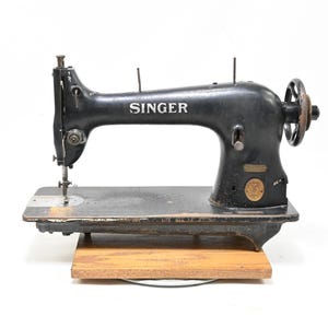 May include: A vintage black Singer sewing machine with a metallic finish, featuring the brand name printed in white on the side, a hand wheel on the right, a needle assembly on the left, and a circular gold emblem, all mounted on a wooden base.