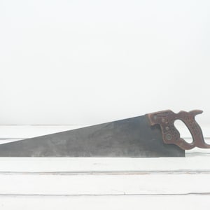 Può includere: Una sega a mano vintage con manico in legno. La sega è in metallo e ha una finitura nera. Il manico è marrone e ha un design decorativo.