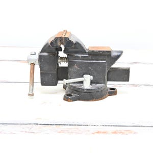 May include: A black and brown bench vice, a tool used to hold workpieces. The vice has a handle, a screw mechanism, and a base for mounting. The jaws are made of metal and the handle is silver. The vice is designed for woodworking or metalworking.