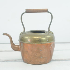 May include: A small copper and brass teapot with a wooden handle. The teapot has a rounded body and a spout on the side.