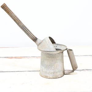 Puede incluir: Aceitera de metal vintage con un pico largo y curvado y un asa. La aceitera es de color plateado mate con un asa y pico texturizados. Tiene un cuerpo cilíndrico y un asa pequeña en el lateral.