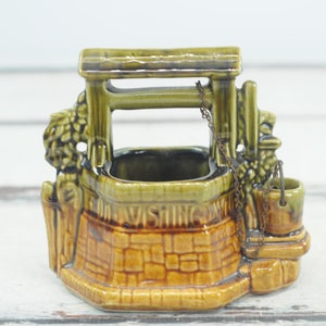 Puede incluir: Un macetero de cerámica con forma de pozo con una base de ladrillo marrón y un marco de madera verde. El pozo tiene una cadena y un cubo, y las palabras "FOR VISITING FRIENDS" están inscritas en el lateral.