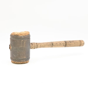 Puede incluir: Un mazo vintage con una cabeza cilíndrica de metal y un mango de madera. La cabeza es de metal oscuro envejecido con un material similar al corcho en la superficie de golpeo. El mango es de madera clara con un agarre estriado. El mazo está sobre un fondo blanco.