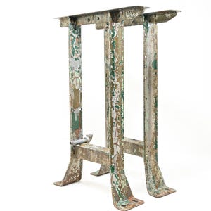 May include: A pair of industrial metal table legs featuring a weathered, distressed finish with patches of peeling green and silver paint. The legs have a vertical H-frame structure with flat mounting plates at the top and wide, flared feet.