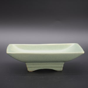 Puede incluir: Un plato rectangular de cerámica verde claro con cuatro patas cortas. El plato está esmaltado y tiene una superficie ligeramente texturizada.
