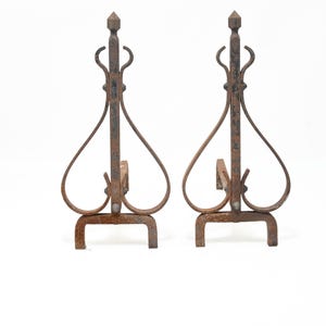 May include: A pair of matching dark brown iron fireplace andirons featuring a vertical central post topped with a pointed finial, flanked by curved metal scrolls that form a heart-like shape, supported by a sturdy rectangular base stand.