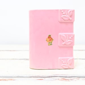 May include: A pink ceramic vase with a rectangular shape. The vase features three decorative square accents with leaf and berry designs. A small floral decal is centered on the front. The vase is set against a white background.